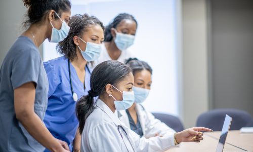 A group of female doctors and nurses are in a meeting together. They are all looking at a laptop screen at the table.